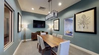 This thoughtfully designed single-floor space functions as an elegant meeting or private dining room, offering a refined environment suited for collaborative work or intimate gatherings. The room is anchored by a rich wooden conference table surrounded by upholstered side chairs in a neutral linen tone, providing both comfort and a polished aesthetic.

The walls are finished in a soothing muted teal, lending the space a calm, professional ambiance. A striking geometric rectangular pendant light in a dark metal finish hangs above the table, serving as a focal point and complementing the room's contemporary design sensibility. Recessed lighting throughout the ceiling ensures the space is evenly and warmly lit.

A wall-mounted flat-screen television is centrally positioned on one wall, making the room well-equipped for presentations or media viewing. Flanking the TV are two tall floor lamps with warm-toned shades, adding layered lighting and a touch of sophistication. A wooden sideboard with decorative cabinet doors sits beneath the TV, offering additional storage and surface space.

Framed artwork adorns the walls, contributing to a curated, gallery-like feel. A window provides a glimpse of an adjacent lounge or outdoor area, adding a sense of connection to the broader environment. The carpeted flooring in a soft gray completes the space with warmth and acoustic comfort.