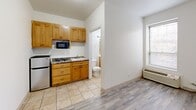 This 180 square foot studio apartment in Chicago offers a well-designed space that maximizes functionality in a compact footprint. The main living area features durable wood-look flooring, creating a warm and inviting atmosphere. A strategically placed kitchenette includes honey-toned wooden cabinetry, providing ample storage, and essential appliances such as a microwave and mini-refrigerator. The bathroom boasts a full-sized tub with a shower, tiled walls in a neutral palette, and a wooden vanity that echoes the kitchen's aesthetic. Large windows allow natural light to flood the space, enhancing the sense of openness. The apartment's layout cleverly separates the sleeping area from the main living space, offering a semblance of distinct zones within the studio. Recessed lighting throughout ensures even illumination, while the neutral color scheme allows for personal customization. Despite its small size, this apartment efficiently accommodates all necessary amenities for comfortable city living, making it an ideal option for those seeking a minimalist lifestyle in an urban setting.