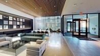This single-floor amenity and lobby space showcases a high-end contemporary design that immediately impresses upon entry. The main lobby is anchored by a striking wood slat ceiling that runs throughout, lending warmth and texture to the otherwise crisp, modern interior. A sleek linear fireplace set into a dark feature wall creates a compelling focal point, while designer lounge seating in deep teal tones, including sculptural chairs and plush sofas, invites residents to linger in comfort. A long white communal bar-height counter with coordinating stools doubles as a gathering and workspace area, complemented by recessed and pendant lighting that adds a refined atmosphere. Floor-to-ceiling glazing and vertical slat screening flood the space with natural light, casting dramatic shadow patterns that shift throughout the day. Adjacent to the lobby, a well-appointed fitness center is enclosed behind sleek black-framed glass partitions, offering visibility and accessibility. Smooth stone-toned accent pieces and a warm wood slat accent wall frame the gym entrance. A dedicated mailroom features an extensive bank of individual mailboxes along polished corridor walls with a clean, functional layout. Stainless steel elevator doors provide seamless access throughout the building. The polished concrete floors unify each zone, contributing to a cohesive and sophisticated atmosphere throughout.
