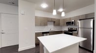 This single-floor residence showcases a clean, modern design throughout. The kitchen is a standout feature, outfitted with flat-front cabinetry in a warm wood-grain finish, crisp white subway tile backsplash, and a full suite of stainless steel appliances including a refrigerator, electric range, built-in microwave, and dishwasher. White quartz countertops extend across the workspace, and a kitchen island with pendant lighting above adds both function and style. Dark hardwood-style flooring runs throughout the main living areas, providing a rich contrast to the light-toned walls. The open-concept layout creates a seamless flow from the entry to the kitchen and living spaces, allowing natural light to travel freely through the unit. A bright bedroom features two windows with views of lush greenery, offering a calm, natural outlook. A carpeted room with a ceiling fan and double French doors opens to an outdoor area, ideal as a second bedroom or flexible living space. The bathroom is well-appointed with a wood-grain vanity, white quartz countertop, a full tub-shower combination, and polished brushed nickel fixtures. The overall aesthetic is contemporary and cohesive, with consistent finishes that lend the space a fresh, move-in-ready appeal.