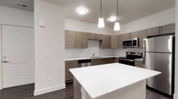 This single-floor residence showcases a clean, modern design throughout. The kitchen is a standout feature, outfitted with flat-front cabinetry in a warm wood-grain finish, crisp white subway tile backsplash, and a full suite of stainless steel appliances including a refrigerator, electric range, built-in microwave, and dishwasher. White quartz countertops extend across the workspace, and a kitchen island with pendant lighting above adds both function and style. Dark hardwood-style flooring runs throughout the main living areas, providing a rich contrast to the light-toned walls. The open-concept layout creates a seamless flow from the entry to the kitchen and living spaces, allowing natural light to travel freely through the unit. A bright bedroom features two windows with views of lush greenery, offering a calm, natural outlook. A carpeted room with a ceiling fan and double French doors opens to an outdoor area, ideal as a second bedroom or flexible living space. The bathroom is well-appointed with a wood-grain vanity, white quartz countertop, a full tub-shower combination, and polished brushed nickel fixtures. The overall aesthetic is contemporary and cohesive, with consistent finishes that lend the space a fresh, move-in-ready appeal.