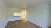 This single-floor space features an open-concept layout that connects the living area to the kitchen, creating a functional and cohesive floor plan. The living area is finished with neutral-toned carpet, providing a comfortable and understated foundation for furnishings. White walls throughout contribute to a clean, bright aesthetic that enhances the sense of openness.

The kitchen is positioned toward the rear of the main living space and is defined by a raised breakfast bar that serves as a natural divider between the two areas. White cabinetry lines the kitchen walls, offering ample storage, while a countertop sink and light-toned counters add to the utilitarian design. A doorway visible from the living area leads further into the unit, suggesting additional rooms beyond the main open space.

Overhead, a ceiling-mounted HVAC vent is visible, indicating a central air system. The flooring transitions from carpet in the living area to a hard surface in the kitchen, clearly delineating the two functional zones. Overall, the space presents a straightforward and practical layout with a neutral palette suited to a variety of uses.