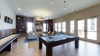 Located at 1150 Helen Drive in Newark, Delaware, this single-floor amenity space is thoughtfully designed to offer residents a sophisticated and inviting retreat. The open-concept layout centers around a full-size pool table with a sleek dark wood frame and blue felt surface, making it an immediate focal point upon entry. Herringbone-patterned hardwood flooring adds a refined architectural touch that flows throughout the space, complementing the warm, neutral color palette accented by deep purple feature walls.

A built-in fireplace with wood-panel surround serves as an elegant anchor to the lounge seating area, which is furnished with plush sofas and accent chairs arranged for comfortable conversation. Flanking the fireplace are floor-to-ceiling built-in shelving units that provide both decorative display space and practical storage. Overhead, a mix of globe pendant lights and a geometric hexagonal chandelier deliver layered, ambient lighting that enhances the room's modern aesthetic.

Abundant natural light pours in through large windows along multiple walls, creating a bright and airy atmosphere while offering views of the surrounding exterior. The combination of stylish finishes, versatile furnishings, and recreational features makes this space well-suited for socializing and leisure.