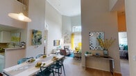 Located at 8720 Costa Verde Blvd in San Diego, the Newport Loft model is a two-story residence that showcases a bold, contemporary design with a vibrant color palette of teal, yellow, and neutral tones throughout. The main living area immediately impresses with dramatically soaring ceilings, crown molding, and an abundance of natural light streaming through a large upper clerestory window and sliding glass doors that open to a balcony. A sleek fireplace serves as a focal point, flanked by a wall-mounted TV and modern media console. The open layout connects the living area to a dedicated dining space, ideal for entertaining. The kitchen features white cabinetry, ample counter space, and hardwood flooring, offering both functionality and style. Two well-appointed bathrooms include tiled tub-shower combinations, hardwood floors, and large vanity mirrors. The bedrooms provide comfortable retreats, with one featuring a bold teal accent wall and another with soft neutral tones, mirrored closet doors, and generous natural light. A second-floor lounge area offers additional flexible living space, complete with high ceilings and curated artwork. Throughout the home, thoughtful design details and colorful accents create a lively yet refined ambiance.