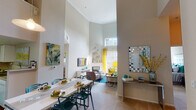 Located at 8720 Costa Verde Blvd in San Diego, the Newport Loft model is a two-story residence that showcases a bold, contemporary design with a vibrant color palette of teal, yellow, and neutral tones throughout. The main living area immediately impresses with dramatically soaring ceilings, crown molding, and an abundance of natural light streaming through a large upper clerestory window and sliding glass doors that open to a balcony. A sleek fireplace serves as a focal point, flanked by a wall-mounted TV and modern media console. The open layout connects the living area to a dedicated dining space, ideal for entertaining. The kitchen features white cabinetry, ample counter space, and hardwood flooring, offering both functionality and style. Two well-appointed bathrooms include tiled tub-shower combinations, hardwood floors, and large vanity mirrors. The bedrooms provide comfortable retreats, with one featuring a bold teal accent wall and another with soft neutral tones, mirrored closet doors, and generous natural light. A second-floor lounge area offers additional flexible living space, complete with high ceilings and curated artwork. Throughout the home, thoughtful design details and colorful accents create a lively yet refined ambiance.