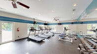 This fitness center at 3006 Hickory Woods Dr offers a comprehensive workout facility designed to support a wide range of exercise routines. The main workout area is generously sized, featuring durable rubber flooring, recessed lighting, and dual ceiling fans that maintain air circulation throughout. Distinctive blue horizontal striping accents the neutral walls, lending the space a clean, energetic aesthetic.

The cardio zone is equipped with multiple treadmills and elliptical machines, positioned along window-lined walls that provide natural light and views of an outdoor pool area. Glass doors on one side offer direct access to the exterior, further enhancing the connection to the outdoors. Ceiling-mounted flat-screen TVs are positioned for easy viewing during cardio sessions.

The strength training area features a full dumbbell rack, weight benches, stability balls, and stationary cycling bikes, complemented by a large floor-to-ceiling mirror displaying the motivational phrase "Your Only Limit Is You." Wall-mounted coat hooks provide convenient storage near the entry.

A dedicated bathroom supports the facility, finished with light blue walls, a wall-mounted sink, mirror, storage cabinetry, and coat hooks — offering practical amenities for post-workout use. Overall, the space delivers a functional and well-appointed fitness environment.