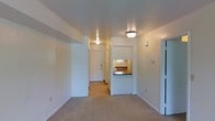 This single-floor space features a straightforward, functional layout with clean lines and a neutral color palette of white and warm beige tones throughout. The main living area is finished with wall-to-wall carpet and smooth, flat ceilings with flush-mount light fixtures providing overhead illumination. A defining architectural detail is the pass-through window connecting the kitchen to the living area, framed by a dark countertop ledge. This opening allows a glimpse into the kitchen, where wood-toned cabinetry and a window providing natural light are visible. A hallway extends toward the rear of the unit, leading to an exterior door, suggesting a secondary entrance or utility area. The transition between the carpeted living space and the hard-surface flooring of the hallway creates a clear functional separation within the layout. White walls are consistent throughout, contributing to a bright, open feel. The overall ambiance is clean and minimal, offering a neutral canvas well-suited for a variety of uses and personal styles.