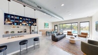 Located at 1418 Frontier Valley Dr in Austin, TX, this single-floor community lounge space offers a thoughtfully designed open-plan layout combining a fully equipped kitchen and a spacious living area. The interior is defined by polished concrete floors, crisp white walls, and an exposed industrial-style HVAC duct that runs prominently across the ceiling, lending the space a modern, loft-inspired character complemented by recessed lighting and pendant fixtures over the kitchen island.

The kitchen is outfitted with light gray shaker cabinetry, a white quartz countertop island with bar seating, a stainless steel refrigerator, a tile backsplash, and a dedicated coffee station. A striking floor-to-ceiling navy blue open shelving unit adds a bold design accent and provides generous display storage.

The living area is generously appointed with a channeled gray sofa, leather accent chairs, a round stone coffee table, and a wall-mounted flat-screen television. A dining table with teal chairs bridges the kitchen and lounge zones seamlessly.

Floor-to-ceiling sliding glass doors and large windows flood the space with natural light and provide direct views of an outdoor pool and landscaped surroundings, creating a bright, inviting ambiance that connects the interior to the outdoors effortlessly.