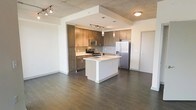 This single-floor space showcases a contemporary open-concept layout that seamlessly integrates the kitchen and living areas. The kitchen is a standout feature, offering warm medium-toned wood cabinetry that extends to the ceiling, providing generous storage. A white kitchen island serves as both a functional workspace and a natural dividing point between the kitchen and the open living area, complete with an undermount sink. Stainless steel appliances, including a french door refrigerator, electric range, and built-in microwave, lend a polished, modern feel. A subway tile backsplash adds a clean, classic touch behind the range. Track lighting illuminates the kitchen area, while a concrete-style ceiling adds an industrial character to the space. Rich, dark hardwood-style flooring runs throughout, grounding the open layout with warmth and continuity. The generous open floor plan between the kitchen and living area creates a spacious, airy atmosphere, ideal for both everyday living and entertaining. The neutral white walls complement the contrasting dark floors and cabinetry tones throughout.