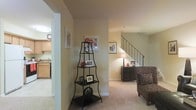 This two-story home offers a comfortable and well-organized layout that balances functionality with a warm, inviting ambiance. The main level features a carpeted living area bathed in soft, ambient light, creating a cozy atmosphere ideal for relaxing or entertaining. A decorative wrought-iron corner shelf adds a stylish accent, while framed artwork along the walls contributes a personalized, homey feel. A staircase with a classic metal railing connects the two floors, serving as both a practical and architectural feature that adds vertical interest to the space. Adjacent to the living area, the kitchen is visible through an open passage, revealing light wood cabinetry and a functional countertop layout that keeps the cooking space accessible and bright. A dresser with a table lamp and additional framed pieces near the staircase provide warm, layered lighting and decorative continuity throughout the main floor. The neutral wall tones and carpeted flooring create a cohesive, understated palette that allows furnishings and décor to stand out. The overall design of the home feels approachable and livable, offering ample space across both floors to accommodate a variety of lifestyle needs.
