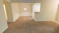 This single-floor residence offers a clean, functional layout designed for comfortable everyday living. The main living and dining area is generously sized, finished with soft, neutral-toned wall-to-wall carpeting that lends a warm and inviting atmosphere throughout the space. A subtle architectural detail divides the dining area from the living space, with a classic cone-shaped pendant light hanging overhead to define the dining zone.

One of the standout features is the pass-through kitchen window, which opens directly into the living area, creating a seamless connection between the kitchen and the main living spaces. The kitchen is fitted with crisp white cabinetry that extends to the upper walls, providing ample storage while contributing to the bright, airy feel of the home. The pass-through counter also offers a convenient breakfast bar or serving surface.

The walls throughout are finished in a soft, neutral tone that enhances the sense of openness and allows for versatile interior styling. Clean white baseboards and trim provide a crisp contrast to the carpeted floors. The overall ambiance is calm and understated, offering a blank canvas well-suited for a variety of furnishing styles. The single-floor layout ensures easy accessibility and a cohesive flow throughout the entire living space.