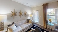 This single-floor residence showcases a cohesive and sophisticated black, gold, and neutral design aesthetic carried throughout every room. The kitchen is outfitted with stainless steel appliances, dark espresso cabinetry, granite countertops, and a double-basin sink, all complemented by pendant lighting and track lighting overhead. Crown molding adds a refined architectural detail that continues into the adjoining spaces. The dining area features a statement chandelier, a circular dining table with upholstered seating, and bold abstract artwork framed in gold, creating an intimate yet elegant setting. The living room offers hardwood-style flooring, a tufted sofa, decorative gold starburst wall accents, crown molding, and access to a private exterior door with natural light filtering through the window. The bedroom is a well-appointed retreat with a tufted upholstered headboard, coordinating nightstands, a ceiling fan, and additional natural light from a window dressed in dramatic dark curtains. The bathroom includes a soaking tub with a curved shower rod, tiled surround, dark vanity cabinetry, and granite countertops, maintaining the space's polished, design-forward character throughout.