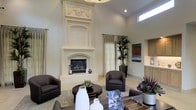 This beautifully appointed single-floor space makes an immediate impression with its soaring ceilings and open, airy atmosphere. The centerpiece of the main living area is a dramatic, floor-to-ceiling ornate fireplace surround crafted in a creamy white finish, featuring intricate carved detailing including decorative drapery motifs at the crown — a true architectural statement piece that anchors the room with classic elegance.

The living area is thoughtfully arranged with dark chocolate-toned upholstered armchairs and a rich, round dark wood coffee table, offering a sophisticated contrast against the soft, neutral wall tones and light flooring. Natural light filters in through tall, sheer linen drapery panels and elevated clerestory windows, lending the space a bright yet serene quality throughout the day.

Lush potted tropical plants introduce organic texture and vibrant greenery, complementing the refined décor. A glimpse into an adjacent area reveals light wood cabinetry, hinting at a well-appointed kitchen or bar space nearby. Colorful artwork adds visual interest to the walls, reinforcing the space's curated, design-forward character. Overall, this space balances grand architectural details with warm, livable comfort.