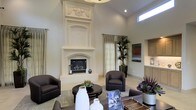 This beautifully appointed single-floor space makes an immediate impression with its soaring ceilings and open, airy atmosphere. The centerpiece of the main living area is a dramatic, floor-to-ceiling ornate fireplace surround crafted in a creamy white finish, featuring intricate carved detailing including decorative drapery motifs at the crown — a true architectural statement piece that anchors the room with classic elegance.

The living area is thoughtfully arranged with dark chocolate-toned upholstered armchairs and a rich, round dark wood coffee table, offering a sophisticated contrast against the soft, neutral wall tones and light flooring. Natural light filters in through tall, sheer linen drapery panels and elevated clerestory windows, lending the space a bright yet serene quality throughout the day.

Lush potted tropical plants introduce organic texture and vibrant greenery, complementing the refined décor. A glimpse into an adjacent area reveals light wood cabinetry, hinting at a well-appointed kitchen or bar space nearby. Colorful artwork adds visual interest to the walls, reinforcing the space's curated, design-forward character. Overall, this space balances grand architectural details with warm, livable comfort.