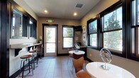 This thoughtfully designed single-floor space blends functionality with contemporary style, making it well-suited as a leasing office, business center, or community amenity space. The interior is finished with neutral-toned walls, large-format light tile flooring, and bold dark-trimmed windows and doors that frame views of surrounding trees, filling the space with natural light throughout.

The main area features multiple seating arrangements, including round white bistro tables paired with warm wood-toned chairs, as well as counter-height bar seating along a narrow wall-mounted ledge near the entrance. A dedicated workstation area is equipped with a built-in granite-topped desk, a printer, and a wall-mounted flat-screen television, supported by framed botanical artwork above.

A coffee and refreshment bar runs along one wall, featuring dark cabinetry, a granite countertop, a Keurig coffee maker, and teal bar stools beneath. Three vibrant framed agate-inspired prints add a pop of color above the bar. A glass-panel interior door provides a view into an adjacent reception or service area, enhancing the open, connected feel of the space. Emergency exit signage and recessed lighting complete the practical yet polished ambiance.