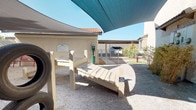 This single-level outdoor dog park is thoughtfully designed to provide a comfortable and functional space for pets and their owners. The area features a gravel ground surface that keeps the space low-maintenance and easy to navigate. Large shade sails stretch overhead, offering relief from direct sunlight and creating a cooler, more comfortable environment during warmer hours. Several slatted lounge-style benches are positioned throughout the space, giving pet owners a place to relax while supervising their animals. A dedicated pet waste station is conveniently located near the entrance, promoting cleanliness and responsible pet ownership. A standout feature of the park is a classic red fire hydrant installed atop a circular patch of artificial turf, adding a whimsical and pet-friendly touch to the design. Ornamental grasses and drought-tolerant plantings line the perimeter walls, introducing natural texture and color to the space. The area is enclosed by chain-link fencing, ensuring pets remain safely contained. Stucco walls with a neutral palette surround the park, consistent with a Southwestern architectural style. Informational signage is posted near the entrance, providing guidelines for park use. Overall, the space balances practicality with thoughtful amenities for both dogs and their owners.