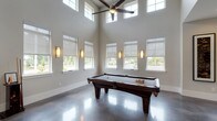 This game room is designed to accommodate a variety of recreational activities within a single, well-appointed space. The room's most striking architectural feature is its soaring double-height ceiling in the billiards area, lined with clerestory windows that flood the space with abundant natural light. A large ceiling fan anchors this section, enhancing both ventilation and visual impact.

The polished concrete flooring runs throughout, providing a sleek, durable surface that ties the entire space together. The billiards zone centers around a traditional dark wood pool table, surrounded by ample space for play. Adjacent to it, a full-length shuffleboard table offers an additional gaming option, accompanied by a wall-mounted flat-screen television and framed artwork.

A dedicated lounge area features a pair of leather chairs, a media console, and a wall-mounted television, creating a comfortable retreat for relaxation. A separate dining table with seating provides space for tabletop games, as evidenced by a tower-stacking game on display. Roller shades on the windows allow for adjustable light control throughout. Wall sconces and recessed lighting complement the natural illumination, creating a well-balanced and inviting atmosphere throughout the room.