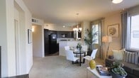 This single-floor residence showcases an open-concept floor plan that integrates the living, dining, and kitchen areas into one cohesive, flowing space. The layout promotes an airy, connected feel, allowing natural light from the dining area's window — dressed with neutral drapes — to filter throughout the space.

The kitchen is equipped with dark cabinetry, black appliances including a refrigerator and built-in microwave, and a light-toned countertop that extends into a breakfast bar. Track lighting runs along the ceiling, supplementing the kitchen's functionality. A chandelier with warm-toned bulbs hangs above the dining area, anchoring a round dark-wood dining table surrounded by contemporary white chairs.

The living area transitions smoothly from the dining space, featuring neutral-toned carpet that adds warmth underfoot. Tall indoor plants introduce organic texture and complement the neutral beige wall tones throughout. The overall ambiance is modern yet approachable, with a cohesive color palette of dark browns, whites, and warm neutrals creating a balanced, inviting atmosphere.