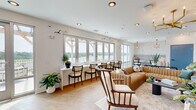Located at 1020 Port Royal Trail in Myrtle Beach, SC, this single-floor coastal space offers a thoughtfully designed lounge and kitchenette area that blends relaxed sophistication with beachside charm. The main lounge area is anchored by a row of large, multi-panel windows that flood the space with natural light and frame sweeping outdoor views of the surrounding landscape. A tufted caramel leather sectional sofa, layered area rugs, and an eclectic mix of accent chairs create a welcoming and stylish seating arrangement. A sage green paneled accent wall serves as a focal point, adorned with a wall-mounted television and topped by a striking gold sputnik chandelier, adding a mid-century modern touch to the coastal aesthetic. Decorative seagull wall sculptures carry the coastal theme throughout. The kitchenette is equally impressive, featuring warm wood cabinetry, quartz countertops, and a bold marble-look tile backsplash and floor. Stainless steel appliances — including a French door refrigerator and double oven — are complemented by a Moravian star pendant light and a deep single-basin sink with a matte black faucet. The seamless transition between wood-look plank flooring in the lounge and hexagonal tile in the kitchen adds visual interest and defines each zone within the open layout.