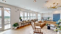 Located at 1020 Port Royal Trail in Myrtle Beach, SC, this single-floor coastal space offers a thoughtfully designed lounge and kitchenette area that blends relaxed sophistication with beachside charm. The main lounge area is anchored by a row of large, multi-panel windows that flood the space with natural light and frame sweeping outdoor views of the surrounding landscape. A tufted caramel leather sectional sofa, layered area rugs, and an eclectic mix of accent chairs create a welcoming and stylish seating arrangement. A sage green paneled accent wall serves as a focal point, adorned with a wall-mounted television and topped by a striking gold sputnik chandelier, adding a mid-century modern touch to the coastal aesthetic. Decorative seagull wall sculptures carry the coastal theme throughout. The kitchenette is equally impressive, featuring warm wood cabinetry, quartz countertops, and a bold marble-look tile backsplash and floor. Stainless steel appliances — including a French door refrigerator and double oven — are complemented by a Moravian star pendant light and a deep single-basin sink with a matte black faucet. The seamless transition between wood-look plank flooring in the lounge and hexagonal tile in the kitchen adds visual interest and defines each zone within the open layout.