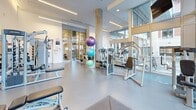 Located at 5121 Bishop St in Halifax, NS, this fitness room offers a well-appointed exercise space designed to support a wide range of workout routines. The room features soaring ceilings and floor-to-ceiling windows along one wall, flooding the space with abundant natural light and providing views of the surrounding urban environment. Exposed concrete columns add an industrial architectural character, complementing the clean, modern aesthetic of white walls and smooth grey flooring throughout.

The exercise area is comprehensively equipped to accommodate diverse fitness needs. A dedicated cardio zone houses a row of treadmills and a rowing machine, positioned to take advantage of the generous natural light. Strength training is well represented, with a selection of cable and weight machines, adjustable benches, and a cable crossover unit. A free weights area features a full rack of dumbbells, kettlebells, and stability balls in multiple sizes, supported by rubber matting for safe floor-based exercises. Ceiling-mounted televisions are positioned throughout for convenience.

Large mirrors enhance the sense of openness and allow for form monitoring during workouts. The space is organized to allow clear circulation between zones, creating a functional and comfortable environment. Informational signage is displayed near the entrance, and hand sanitizing stations are conveniently accessible, reflecting a focus on cleanliness and user well-being.