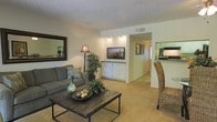 This single-story residence showcases an open-concept design that seamlessly connects the living area to the kitchen and adjacent spaces. The living room serves as the central gathering space, featuring wall-to-wall carpeting in a neutral beige tone that adds warmth underfoot. A built-in media niche with recessed accent lighting and white cabinetry below provides a dedicated and organized space for entertainment. The room is furnished with a comfortable grey sofa accented with decorative pillows, complemented by a rustic wooden coffee table that adds textural contrast. A large framed mirror and a classic floor lamp contribute to the room's inviting ambiance. Lush indoor plants introduce a natural element, enhancing the overall aesthetic. A wicker accent chair near the kitchen bar area bridges the living and dining spaces. The kitchen is partially visible, featuring bar-style seating and dark cabinetry, suggesting a functional layout ideal for casual dining. A hallway leading to additional rooms is visible, lined with framed artwork. The textured ceiling and neutral wall tones throughout create a cohesive, understated palette across the entire space.