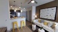 This single-floor residence in Carrollton, Texas, offers a thoughtfully designed open-concept layout that seamlessly blends the kitchen and living areas into one cohesive, functional space. The kitchen is outfitted with dark cabinetry, stainless steel appliances, and a granite-topped breakfast bar with seating — perfect for casual dining or entertaining. Stylish pendant lighting hangs above the counter, complementing the track lighting and adding a modern touch. The living area is warm and welcoming, anchored by a neutral-toned sofa accented with yellow and gray throw pillows that echo the bold yellow bar stools at the kitchen counter. A ceiling fan centers the space, while a rich, dark accent wall provides depth and contrast against the lighter surrounding walls. Wood-look flooring runs throughout, adding warmth and continuity to the open floor plan. A large piece of artwork adorns the accent wall, enhancing the curated aesthetic. Hallway doors lead to additional areas of the home, suggesting a well-organized layout beyond the main living space. The overall ambiance is modern, clean, and vibrant, with a cohesive color palette of yellow, white, gray, and brown that gives the space a lively yet refined character.