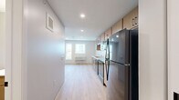 This studio unit at 2945 Garfield Ave in Minneapolis, MN offers a well-organized, efficiently designed single-floor layout. Light wood laminate flooring runs continuously throughout the space, lending a clean, cohesive aesthetic that enhances the sense of openness.

The kitchen is positioned along one wall and features warm-toned wood cabinetry with black hardware, extending both above and below the counter for ample storage. A white countertop provides functional workspace, complemented by a stainless steel sink, black electric range, black dishwasher, and a full-size black refrigerator. A built-in microwave is mounted above the range, completing the fully equipped setup. Recessed lighting illuminates the kitchen area effectively.

The open living area benefits from natural light through a window that offers exterior views of the surrounding neighborhood. Neutral gray walls and clean white ceilings create a bright, understated atmosphere throughout. A wall-mounted HVAC unit provides in-unit climate control.

The bathroom includes a vanity with wood cabinetry matching the kitchen, a sink, toilet, and a dedicated walk-in shower stall with white surround panels and chrome fixtures. A sliding barn-style door with black hardware adds a modern touch to the bathroom entry.

A short entry corridor connects the front door to the main living space, with a sliding closet panel providing additional storage. The unit's straightforward layout maximizes functionality within its compact footprint.