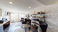 This single-floor space blends industrial character with refined comfort, creating an inviting lounge or common area with a distinctly curated aesthetic. The most striking architectural feature is the expansive whitewashed brick accent wall, which spans the length of the main gathering area and serves as a dramatic backdrop for floor-to-ceiling open shelving in wood and black metal. These shelves are thoughtfully arranged with books, ceramics, framed artwork, and decorative objects, lending the space a lived-in, sophisticated feel.

Rich, dark hardwood flooring runs throughout, anchoring the room and complementing the deep tones of the upholstered wingback and accent chairs arranged around a round pedestal table. A vintage-style area rug defines the central seating zone, adding warmth and texture underfoot. Generous windows with sheer linen drapes flood the space with abundant natural light, framing views of mature trees outside.

The entryway area features a practical built-in credenza with dark cabinetry and a light countertop, paired with frosted glass windows and a bold double-door entry with glass pane detailing. Recessed lighting throughout ensures the space is well-lit at all times. Overall, the space exudes an elegant yet approachable ambiance, combining industrial and transitional design elements seamlessly.