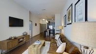 This single-floor space showcases an open-concept layout that connects the living area and kitchen in one continuous flow. Dark hardwood flooring runs throughout, providing a warm, grounding contrast to the neutral, light-toned walls. The living area is anchored by a rich brown leather sofa accented with decorative pillows, paired with a rustic wooden coffee table that reinforces the space's blend of modern and industrial design. A wall-mounted flat-screen TV and a media console with hairpin legs complete the living setup, while framed artwork on the walls adds a curated, personal touch.

The kitchen, visible from the living area, features dark cabinetry and stainless steel appliances, including a full-sized refrigerator. A kitchen island with bar-height seating provides additional dining and prep space, illuminated by pendant lighting overhead. Track lighting is also incorporated, allowing for flexible illumination throughout the open space. A hallway extending from the living area leads to additional rooms, suggesting a thoughtful and functional floor plan. Overall, the space presents a cohesive, contemporary aesthetic with warm, inviting undertones.