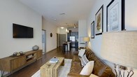 This single-floor space showcases an open-concept layout that connects the living area and kitchen in one continuous flow. Dark hardwood flooring runs throughout, providing a warm, grounding contrast to the neutral, light-toned walls. The living area is anchored by a rich brown leather sofa accented with decorative pillows, paired with a rustic wooden coffee table that reinforces the space's blend of modern and industrial design. A wall-mounted flat-screen TV and a media console with hairpin legs complete the living setup, while framed artwork on the walls adds a curated, personal touch.

The kitchen, visible from the living area, features dark cabinetry and stainless steel appliances, including a full-sized refrigerator. A kitchen island with bar-height seating provides additional dining and prep space, illuminated by pendant lighting overhead. Track lighting is also incorporated, allowing for flexible illumination throughout the open space. A hallway extending from the living area leads to additional rooms, suggesting a thoughtful and functional floor plan. Overall, the space presents a cohesive, contemporary aesthetic with warm, inviting undertones.