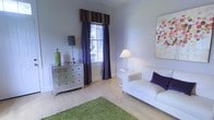 This well-appointed two-story space offers a thoughtfully designed interior that blends comfort with understated elegance. The living area immediately draws attention with its light hardwood flooring, which adds warmth and a sense of openness to the room. Soft, neutral wall tones in a cool lavender-grey hue create a calming backdrop that complements the carefully chosen furnishings, including a clean-lined white sofa accented with a rich purple throw pillow. A mirrored dresser with chrome hardware adds a touch of glamour, while a vibrant green area rug introduces a fresh pop of color that enlivens the space. Natural light filters in through a large window dressed with deep purple silk drapes and a striped fabric valance, providing both style and privacy. A classic table lamp and an additional standing lamp ensure warm, layered lighting throughout the room. Decorative artwork on the walls adds a personal, curated feel to the interior. The overall ambiance is refined yet welcoming, with a cohesive color palette and attention to detail that carries throughout both floors. The space offers a versatile and comfortable environment suited for a variety of uses.