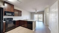 The Piñon floor plan at 5300 Outlook Blvd in Pueblo, Colorado, is a thoughtfully designed one-bedroom, one-bathroom residence on a single level. The open-concept layout seamlessly connects the kitchen and living room, creating a cohesive and functional living space. The kitchen is well-equipped, featuring rich dark-brown cabinetry, granite-style countertops, a peninsula with a built-in sink, and a full suite of black appliances including a refrigerator, range, over-the-range microwave, and dishwasher. Light wood-style flooring runs throughout the unit, complementing the neutral, warm-toned walls. The living room is bright and airy, highlighted by a ceiling fan and a large sliding glass door with vertical blinds that opens to an outdoor patio, allowing generous natural light. The bedroom is a comfortable, quiet retreat with a window offering exterior views and access to a dedicated closet. The bathroom features matching dark cabinetry with granite-style counters, a tub-shower combo with tiled surround, and a walk-in closet conveniently accessible from within. Clean lines, consistent finishes, and a practical layout define this residence's overall character.
