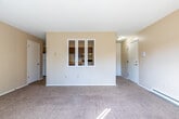 This single-floor unit offers a straightforward, functional layout with a variety of living spaces bathed in natural light. The living area is generously proportioned, featuring neutral beige walls, plush wall-to-wall carpeting, and a full-width sliding glass door fitted with vertical blinds that opens to an outdoor view, allowing abundant sunlight to fill the room. A wall-mounted air conditioning unit and baseboard heating provide year-round climate control.

A second carpeted room, suitable as a bedroom or additional living space, includes a window with blinds overlooking greenery, baseboard heating, and a built-in closet with louvered doors and shelving. An adjacent room also features carpeting and direct sightlines to the kitchen, which is equipped with a white electric range, an under-cabinet range hood, a double-basin stainless steel sink, light wood laminate countertops, and warm-toned wood cabinetry providing ample storage. The kitchen floor is finished in neutral ceramic tile.

The bathroom includes a full tub-shower combination, a single-sink vanity with cabinet storage below, and tile flooring that complements the warm tones found throughout the unit. Overall, the space presents a clean, practical interior with consistent finishes and comfortable natural light.