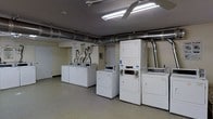 This single-floor laundry facility is designed with functionality and practicality in mind. The room is well-lit by overhead fluorescent lighting, ensuring a bright and comfortable environment for users. Exposed industrial-style ductwork runs along the ceiling, adding a utilitarian character to the space while maintaining efficient ventilation throughout.

The room is equipped with multiple front-loading washing machines and both stackable and standalone dryers, all in white, neatly arranged along the walls and in rows to maximize the use of space. The machines appear to be coin- or card-operated, making this a convenient shared laundry facility. A utility area near the back wall features plumbing connections and informational signage to guide users.

The flooring is a smooth, durable surface suited for high-traffic use, and the neutral-toned walls contribute to the clean, organized aesthetic of the space. A door on the far wall provides additional access or utility. Overall, the layout is efficient and straightforward, offering a practical and well-maintained environment for laundry use.