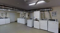 This single-floor laundry facility is designed with functionality and practicality in mind. The room is well-lit by overhead fluorescent lighting, ensuring a bright and comfortable environment for users. Exposed industrial-style ductwork runs along the ceiling, adding a utilitarian character to the space while maintaining efficient ventilation throughout.

The room is equipped with multiple front-loading washing machines and both stackable and standalone dryers, all in white, neatly arranged along the walls and in rows to maximize the use of space. The machines appear to be coin- or card-operated, making this a convenient shared laundry facility. A utility area near the back wall features plumbing connections and informational signage to guide users.

The flooring is a smooth, durable surface suited for high-traffic use, and the neutral-toned walls contribute to the clean, organized aesthetic of the space. A door on the far wall provides additional access or utility. Overall, the layout is efficient and straightforward, offering a practical and well-maintained environment for laundry use.