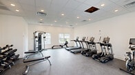 This fitness center in North Augusta, SC, is a well-organized, single-floor facility designed to support a well-rounded workout routine. The space features a drop-panel ceiling with recessed lighting, providing consistent, even illumination throughout. Durable speckled rubber flooring covers the main exercise area, offering both traction and cushioning for high-impact activities.

The cardio section is equipped with multiple TRUE-brand treadmills, ellipticals, and a stationary bike, all arranged in a row along one wall. A rowing machine and a multi-function cable resistance station round out the strength and conditioning equipment. Windows along the interior wall allow natural light to filter into the space, contributing to an open and airy atmosphere.

A separate flexible area near the French door entrance provides additional open floor space, suitable for stretching, yoga, or group exercise. This section includes stability balls and a yoga mat holder. The French doors offer views of the exterior and allow natural light to enhance the brightness of the room. A large interior door provides access to an adjacent storage or utility room. White walls and clean finishes throughout give the fitness center a modern, welcoming appearance.