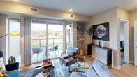 This thoughtfully designed single-floor residence showcases a clean, modern aesthetic throughout its open-concept layout. The kitchen is a standout feature, equipped with sleek wood-grain cabinetry extending to the ceiling, stainless steel appliances including a gas range, built-in microwave, and French door refrigerator, complemented by quartz countertops, a light glass tile backsplash, and a double-basin sink illuminated by stylish pendant lighting. The living area is bright and welcoming, with large sliding glass doors opening to a private balcony that fills the space with natural light and offers open outdoor views. Light hardwood flooring runs throughout the main living areas, adding warmth to the contemporary design. An in-unit stacked washer and dryer is conveniently tucked away near the living space. The primary bedroom features a ceiling fan, a large window with outdoor views, and direct access to a well-appointed bathroom complete with a walk-in glass-enclosed shower with a rainfall showerhead and an adjoining walk-in closet with open shelving. The second bedroom offers a quiet, cozy retreat with soft neutral tones. The overall ambiance of the residence is contemporary and inviting, blending functional design with modern finishes.
