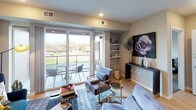 This thoughtfully designed single-floor residence showcases a clean, modern aesthetic throughout its open-concept layout. The kitchen is a standout feature, equipped with sleek wood-grain cabinetry extending to the ceiling, stainless steel appliances including a gas range, built-in microwave, and French door refrigerator, complemented by quartz countertops, a light glass tile backsplash, and a double-basin sink illuminated by stylish pendant lighting. The living area is bright and welcoming, with large sliding glass doors opening to a private balcony that fills the space with natural light and offers open outdoor views. Light hardwood flooring runs throughout the main living areas, adding warmth to the contemporary design. An in-unit stacked washer and dryer is conveniently tucked away near the living space. The primary bedroom features a ceiling fan, a large window with outdoor views, and direct access to a well-appointed bathroom complete with a walk-in glass-enclosed shower with a rainfall showerhead and an adjoining walk-in closet with open shelving. The second bedroom offers a quiet, cozy retreat with soft neutral tones. The overall ambiance of the residence is contemporary and inviting, blending functional design with modern finishes.