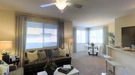 This single-floor residence features an open-concept design that seamlessly integrates the living and dining areas into one cohesive, light-filled space. The living area is anchored by plush wall-to-wall carpeting in a warm neutral tone, providing comfort underfoot and complementing the overall color palette of soft beiges and grays. A ceiling fan with an integrated light fixture serves as a functional centerpiece overhead, ensuring year-round comfort.

Large windows dressed with horizontal blinds and elegant gray curtain panels line the walls, flooding the space with generous natural light while offering adjustable privacy. The living area accommodates a dark upholstered sofa styled with coordinating throw pillows, paired with a sleek white coffee table for a contemporary contrast.

Adjacent to the living area, the dining space features a round glass-top table surrounded by modern white chairs, lit by a chandelier overhead. The open sightlines between both zones enhance the sense of spaciousness, making the layout feel airy and well-connected. The neutral wall tones and consistent flooring unify the space, creating a clean and inviting ambiance throughout.