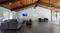 This single-floor space is defined by its striking architectural character, anchored by soaring vaulted ceilings clad in warm, rich wood planking and accented by bold dark exposed beams that run the length of the structure. Dark-toned hardwood-style flooring flows consistently throughout, providing a cohesive and polished aesthetic across all areas.

The expansive main lounge area offers generous seating with multiple gray upholstered sofas and accent chairs arranged around a wall-mounted flat-screen television. A bank of large windows along one wall fills the space with natural light and frames outdoor views. Ceiling fans are integrated into the vaulted ceiling, enhancing comfort throughout.

A reception or check-in area with a white counter divides the main lounge from additional spaces, which include a dedicated game room featuring a ping pong table, pendant lighting, and seating along windowed walls. A separate dining area opens toward what appears to be an outdoor pool area through large glass doors and expansive windows, flooding the room with light.

The fully equipped kitchen features dark cabinetry, granite-style countertops, and black appliances in a functional U-shaped layout. Throughout, the space maintains a modern yet welcoming ambiance suited for community gatherings and events.