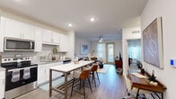 This single-story unit offers a thoughtfully designed open-concept layout that maximizes its compact footprint. The kitchen serves as a central feature, showcasing white shaker cabinetry, granite countertops, a subway tile backsplash, stainless steel appliances including a French door refrigerator and gas range, and a large island with seating — ideal for both meal preparation and casual dining. Hardwood-style flooring runs throughout the main living areas, lending a clean, contemporary aesthetic.

The living room benefits from natural light streaming through full-length windows and a glass door, complemented by a ceiling fan overhead. A dedicated workspace area is conveniently positioned along one wall, adding functional versatility to the open floor plan.

The bedroom features plush carpeting, a ceiling fan, large windows with sheer curtains, and a bold accent wall that adds visual depth to the space. A closet provides organized storage.

The bathroom includes a granite-topped vanity, wood-style flooring, and a shower/tub combination. A utility room and additional closet round out the practical amenities, ensuring efficient use of every area within this well-appointed unit.