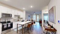 This single-story unit offers a thoughtfully designed open-concept layout that maximizes its compact footprint. The kitchen serves as a central feature, showcasing white shaker cabinetry, granite countertops, a subway tile backsplash, stainless steel appliances including a French door refrigerator and gas range, and a large island with seating — ideal for both meal preparation and casual dining. Hardwood-style flooring runs throughout the main living areas, lending a clean, contemporary aesthetic.

The living room benefits from natural light streaming through full-length windows and a glass door, complemented by a ceiling fan overhead. A dedicated workspace area is conveniently positioned along one wall, adding functional versatility to the open floor plan.

The bedroom features plush carpeting, a ceiling fan, large windows with sheer curtains, and a bold accent wall that adds visual depth to the space. A closet provides organized storage.

The bathroom includes a granite-topped vanity, wood-style flooring, and a shower/tub combination. A utility room and additional closet round out the practical amenities, ensuring efficient use of every area within this well-appointed unit.