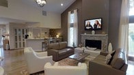This single-floor home showcases a thoughtfully designed open-concept layout that blends comfort and style. The living area immediately draws the eye upward with its dramatic vaulted ceilings, creating a spacious and airy atmosphere. A striking stone-surround fireplace serves as the focal point of the room, complemented by a wall-mounted flat-screen television above it. The seating arrangement features a mix of rich brown leather sofas and cream upholstered accent chairs, anchored by a plush shag area rug that adds warmth and texture to the space. A tall window dressed with elegant white curtains allows natural light to filter in, further enhancing the open feel of the room. An iron chandelier adds a refined decorative touch overhead. The living space flows seamlessly into the kitchen and dining area, which features a raised breakfast bar with wooden counter stools, under-cabinet lighting, and white cabinetry. The overall color palette — warm taupe walls, neutral furnishings, and natural wood accents — creates a cohesive, inviting ambiance throughout. This home strikes a balance between casual livability and sophisticated interior design, making it both functional and visually appealing.