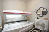 This single-room tanning space is designed with functionality and comfort in mind, offering a clean and well-appointed environment for UV tanning sessions. The centerpiece of the room is a professional-grade tanning bed, featuring a sleek white and red canopy with an array of UV lamps and a built-in digital control panel, ensuring a consistent and controlled tanning experience. The room's neutral warm-beige walls create a calm, inviting atmosphere, while the bold zebra-print area rug adds a stylish, energetic accent underfoot. A white vanity cabinet with a decorative circular-patterned front panel sits against one wall, topped with a few personal care items and accented by an ornate black quatrefoil mirror above. Informational signage, including tanning bed usage guidelines and safety warnings, is prominently posted on the walls, keeping users well-informed. A small waste bin is conveniently placed near the tanning unit. The room is accessed through a solid paneled door, providing privacy during sessions. The overall layout is efficiently organized, maximizing the available space while maintaining a tidy, professional atmosphere suited for a salon or wellness facility.