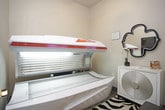 This single-room tanning space is designed with functionality and comfort in mind, offering a clean and well-appointed environment for UV tanning sessions. The centerpiece of the room is a professional-grade tanning bed, featuring a sleek white and red canopy with an array of UV lamps and a built-in digital control panel, ensuring a consistent and controlled tanning experience. The room's neutral warm-beige walls create a calm, inviting atmosphere, while the bold zebra-print area rug adds a stylish, energetic accent underfoot. A white vanity cabinet with a decorative circular-patterned front panel sits against one wall, topped with a few personal care items and accented by an ornate black quatrefoil mirror above. Informational signage, including tanning bed usage guidelines and safety warnings, is prominently posted on the walls, keeping users well-informed. A small waste bin is conveniently placed near the tanning unit. The room is accessed through a solid paneled door, providing privacy during sessions. The overall layout is efficiently organized, maximizing the available space while maintaining a tidy, professional atmosphere suited for a salon or wellness facility.