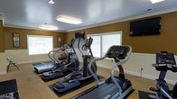 This fitness center offers a dedicated space for cardiovascular and general exercise, thoughtfully arranged to maximize both functionality and comfort. The room is finished with warm golden-toned walls accented by white crown molding, lending the space a clean and polished aesthetic. Overhead, flush-mounted fluorescent light fixtures provide bright, even illumination throughout, while multiple windows fitted with white blinds allow natural light to filter in, creating an inviting workout environment. The flooring features a combination of patterned carpet and protective rubber mats positioned beneath the equipment, ensuring stability and safety during exercise. The cardio equipment lineup includes treadmills and elliptical trainers, catering to a range of fitness preferences and workout intensities. Multiple wall-mounted flat-screen televisions are strategically placed throughout the room, allowing users to stay entertained during their sessions. A stability ball is also visible, indicating additional options for functional training and core work. The space is compact yet efficiently organized, making it easy to navigate between equipment. Overall, this fitness center provides a comfortable, well-lit, and adequately equipped environment suited for residents or guests seeking convenient access to daily exercise facilities.