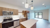 This well-appointed single-floor unit offers a thoughtfully designed open-concept layout that seamlessly connects the kitchen and living areas. The kitchen is a standout feature, showcasing flat-front wood-toned cabinetry, crisp white quartz countertops, and a full suite of stainless steel appliances including a refrigerator, electric range, microwave, and dishwasher. A white subway tile backsplash adds a clean, contemporary touch, while a center island provides additional prep and gathering space. Pendant lighting with globe-style fixtures illuminates the kitchen and extends into the adjacent living area, enhancing the warm ambiance throughout.

The flooring is finished in durable wood-look planks that run consistently across the main living spaces, grounding the design with a natural aesthetic. One of the most notable features is an arched window in a bedroom or flex space that frames a scenic waterfront view, flooding the room with natural light. A sliding glass door in the living area offers access to an outdoor balcony.

The bathroom is clean and functional, featuring a walk-in shower with built-in shelving, a frosted glass door, and neutral tile flooring. The 3D overview reveals a well-organized floorplan with defined spaces for living, cooking, sleeping, and bathing, all within a compact yet efficient footprint.