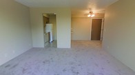 This single-floor space features an open-concept layout that connects the main living area to the kitchen. The living area is finished with wall-to-wall carpet in a neutral tone, creating a soft underfoot feel throughout the space. A ceiling fan with an integrated light fixture is centrally positioned, providing both air circulation and ambient lighting to the room. The entry door is situated along one wall, offering a straightforward and functional entryway.

The kitchen is accessible directly from the living area and is finished with tile flooring, providing a practical contrast to the carpeted living space. Standard kitchen appliances are included, featuring a white refrigerator and a stove with an overhead range, complemented by wooden cabinetry above for storage. The layout is compact yet functional, making efficient use of the available space.

The walls throughout are painted in a consistent light, neutral tone, contributing to a clean and understated aesthetic. The overall design of the space is simple and practical, prioritizing functionality with an open flow between the main living and kitchen areas.