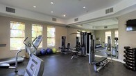 This fitness center offers a comprehensive workout environment designed to accommodate a wide range of exercise routines. The space is finished with durable dark rubber flooring, ideal for high-traffic gym use, and features a clean, neutral color palette with light gray walls and a white ceiling that contributes to a bright, open atmosphere. Recessed lighting is evenly distributed throughout, ensuring the entire area is well-lit during any hour of the day. Large windows along one wall allow natural light to filter in, enhancing the overall ambiance and providing views of the surrounding greenery. A full-length wall mirror on one side of the room visually expands the space and allows users to monitor their form during workouts. The facility is outfitted with a selection of cardio equipment, including treadmills and an elliptical machine, alongside a multi-station cable strength machine and an adjustable workout bench. Exercise balls in multiple sizes are also available for core and flexibility training. Ventilation is well-considered, with multiple ceiling vents ensuring comfortable air circulation. The single-floor layout provides an accessible and straightforward experience, making this fitness center a practical and fully functional facility.
